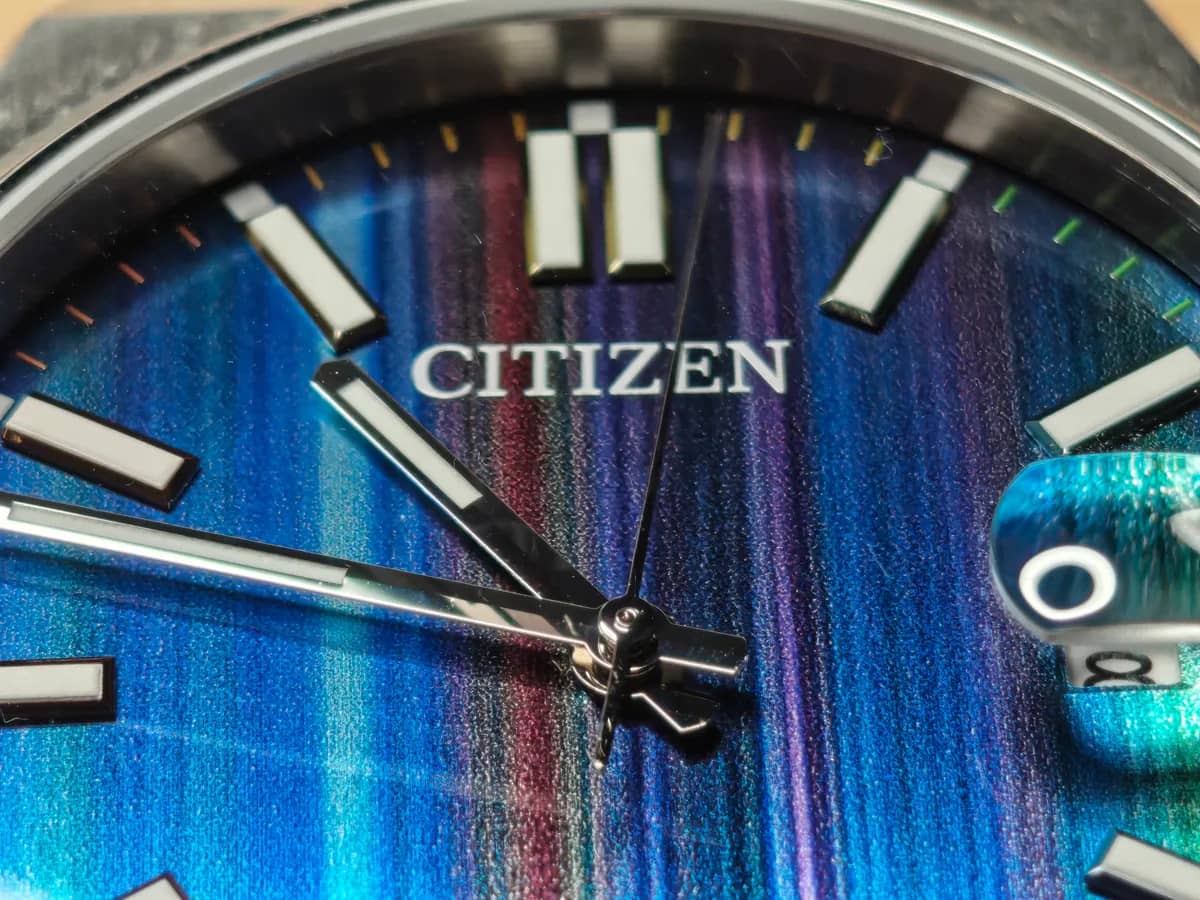 Macro de la esfera: la textura granulada de trazos verticales tipo lápiz de color bajo el logotipo CITIZEN y los índices aplicados con lumen