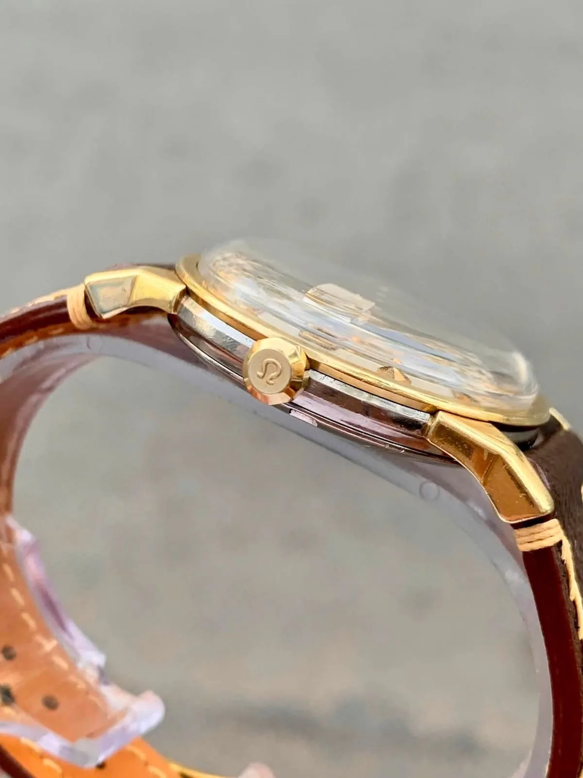 Macro del canto: corona dorada con el logotipo Omega, plexiglás abombado y asa facetada de la caja gold cap