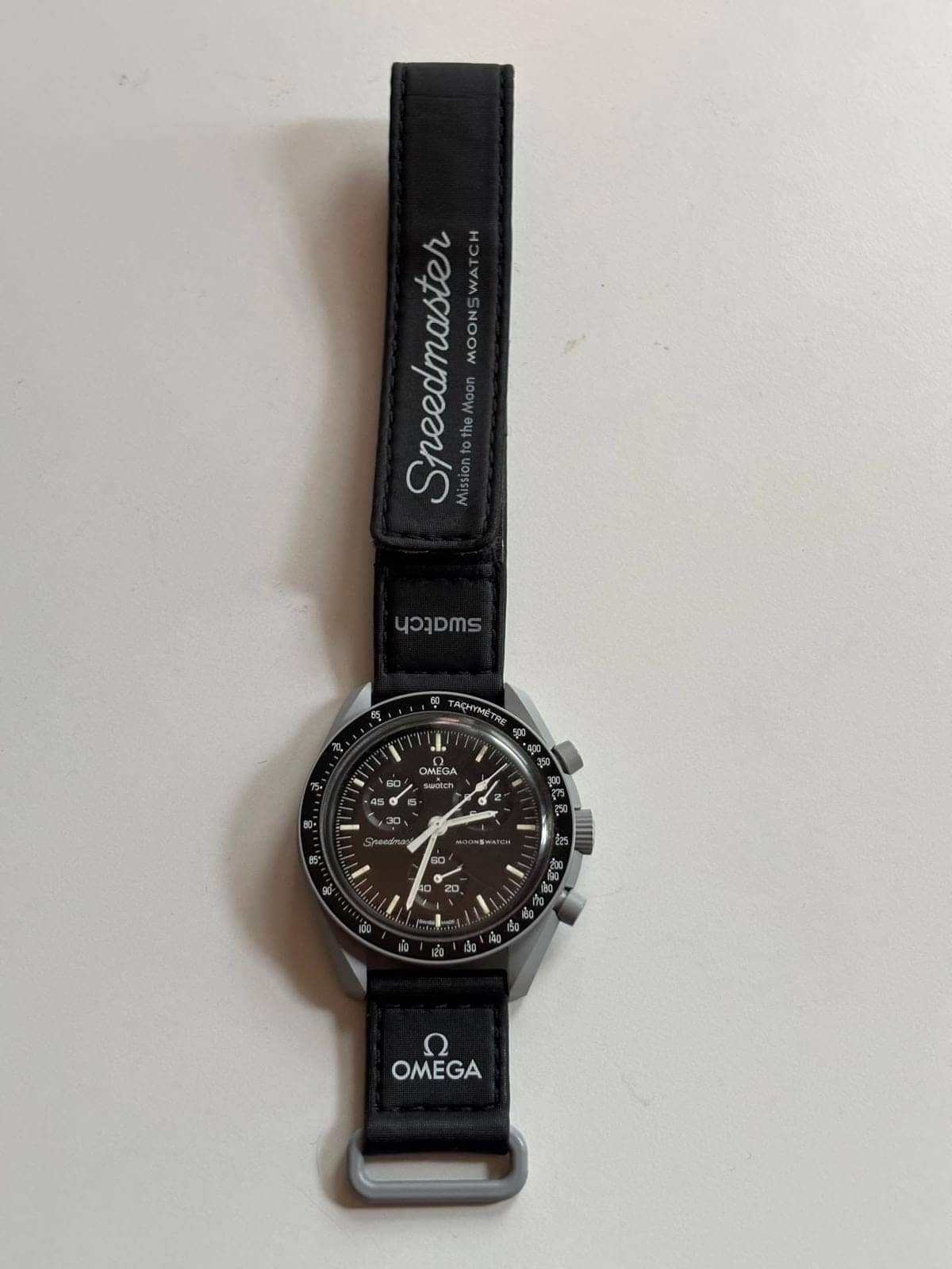 Vista completa del reloj con la correa de velcro extendida, firmada «Speedmaster — Mission to the Moon — MoonSwatch»