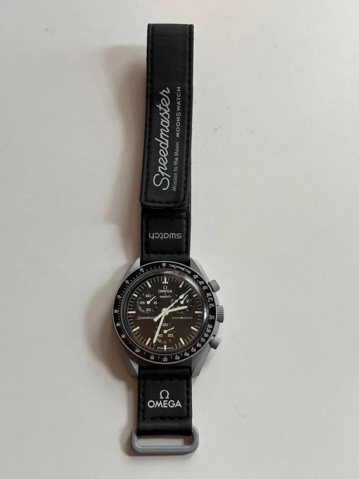 Vista completa del reloj con la correa de velcro extendida, firmada «Speedmaster — Mission to the Moon — MoonSwatch»