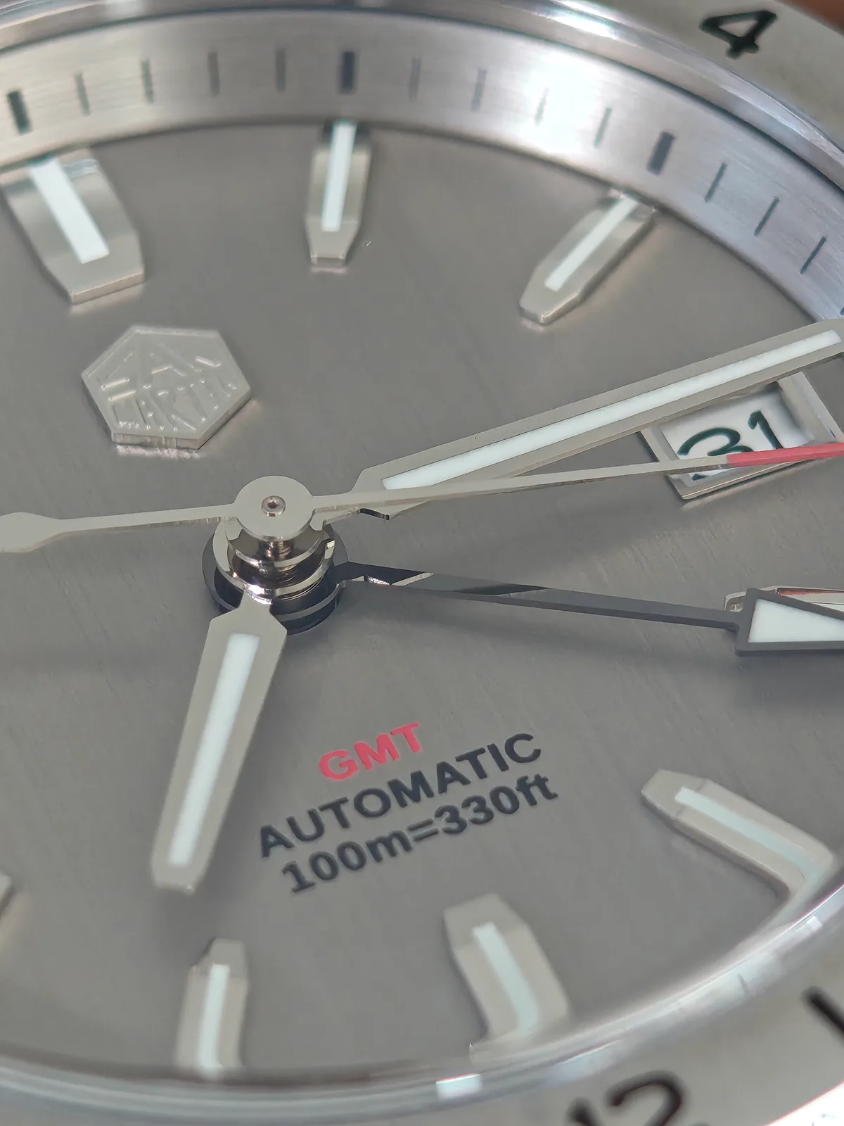 Macro del dial: el rótulo GMT en rojo, las agujas con lumen y la aguja de 24 horas con punta de flecha
