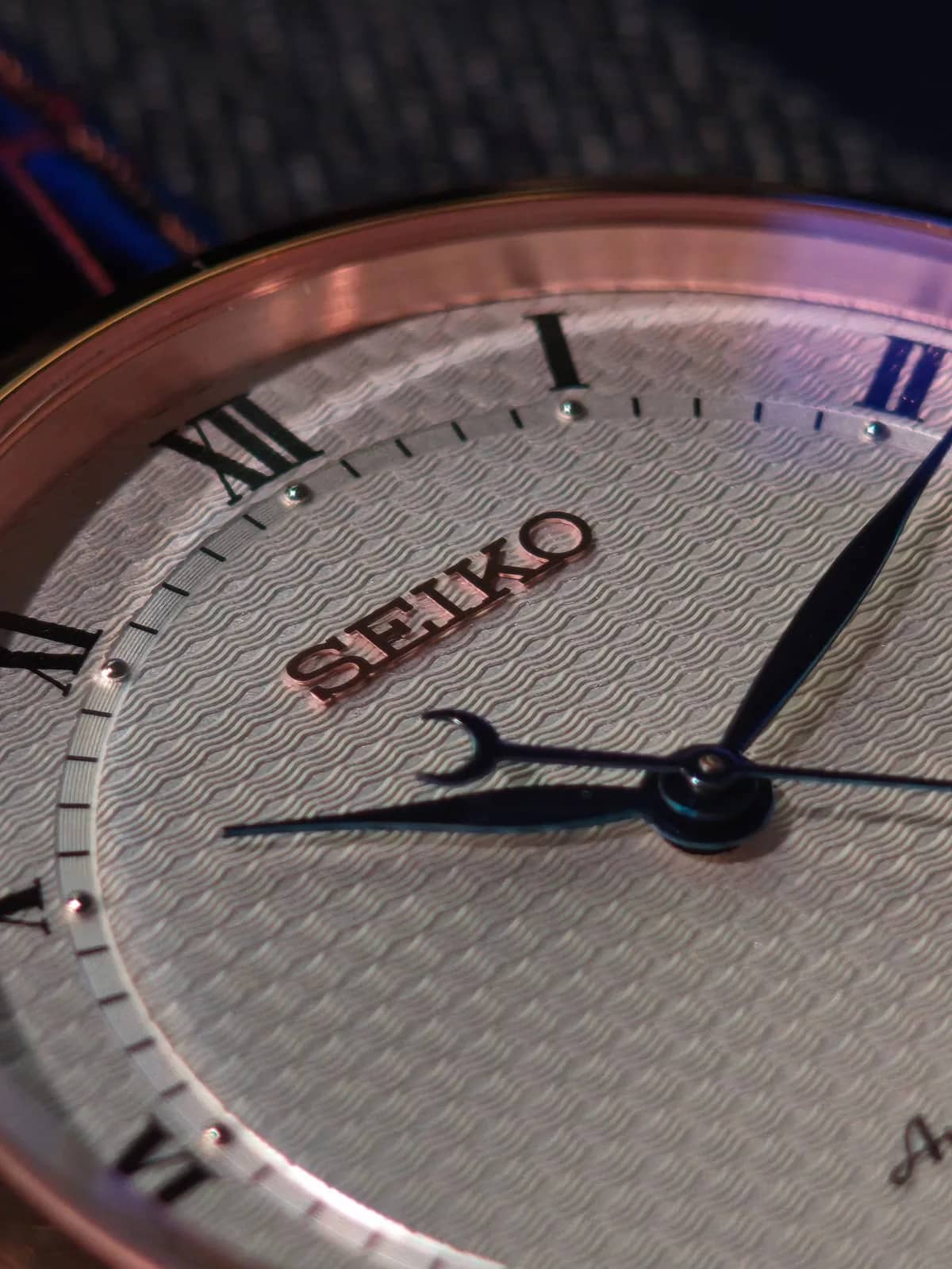 Macro de la esfera: el logotipo SEIKO aplicado en tono dorado sobre el guilloché de ondas y la aguja horaria con contrapeso de media luna