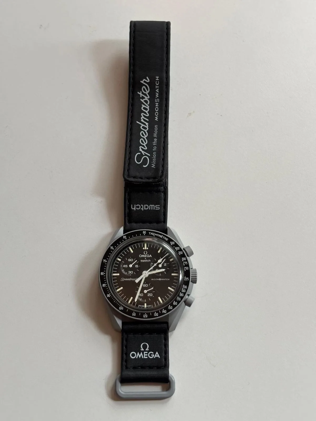 Vista completa del reloj con la correa de velcro extendida, firmada «Speedmaster — Mission to the Moon — MoonSwatch»