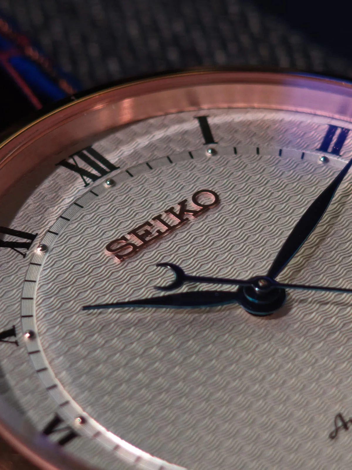 Macro de la carátula: el logotipo SEIKO aplicado en tono dorado sobre el guilloché de ondas y la manecilla horaria con contrapeso de media luna